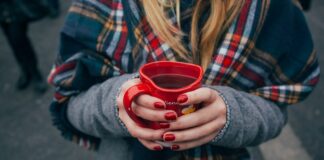 Eine Person mit langen blonden Haaren in Wintermantel mit einem karierten Schal hält ein herzförmiges Punsch-Häferl in Händen mit rot manikürten Nägeln. Fokus ist auf das Punschhäferl, Gesicht und Beine sind nicht zu sehen. Im Hintergrund sind die Beine anderer Leute zu sehen, die auf Asphalt stehen.
