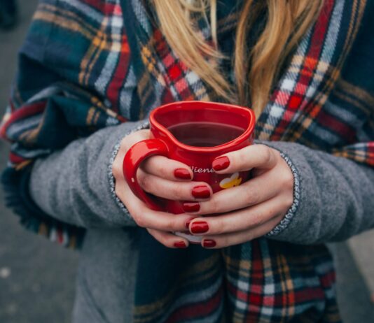Eine Person mit langen blonden Haaren in Wintermantel mit einem karierten Schal hält ein herzförmiges Punsch-Häferl in Händen mit rot manikürten Nägeln. Fokus ist auf das Punschhäferl, Gesicht und Beine sind nicht zu sehen. Im Hintergrund sind die Beine anderer Leute zu sehen, die auf Asphalt stehen.