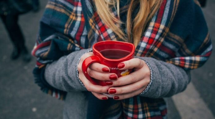 Gefährliche Adventwochen: Mehr Unfälle durch Alkohol im Straßenverkehr Eine Person mit langen blonden Haaren in Wintermantel mit einem karierten Schal hält ein herzförmiges Punsch-Häferl in Händen mit rot manikürten Nägeln. Fokus ist auf das Punschhäferl, Gesicht und Beine sind nicht zu sehen. Im Hintergrund sind die Beine anderer Leute zu sehen, die auf Asphalt stehen.