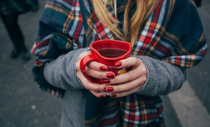 Eine Person mit langen blonden Haaren in Wintermantel mit einem karierten Schal hält ein herzförmiges Punsch-Häferl in Händen mit rot manikürten Nägeln. Fokus ist auf das Punschhäferl, Gesicht und Beine sind nicht zu sehen. Im Hintergrund sind die Beine anderer Leute zu sehen, die auf Asphalt stehen.