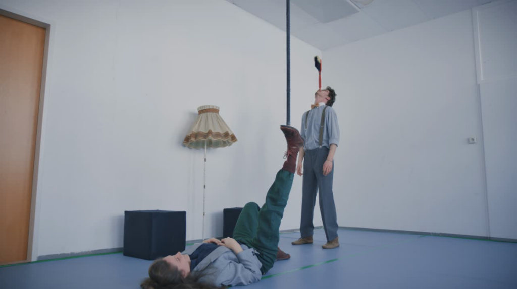 Eine Frau liegt am Boden und balanciert einen Besen-Stiel auf ihrem rechten Fuß. Ein Mann steht daneben und balanciert einen Kehrbesen auf der Nase.