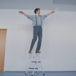 Ein Mann balanciert auf dem Griff einer Trittleiter und hält dabei einen Staubwedel in der Hand.