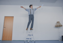 Gefährliche Balanceakte beim Frühjahrsputz Ein Mann balanciert auf dem Griff einer Trittleiter und hält dabei einen Staubwedel in der Hand.