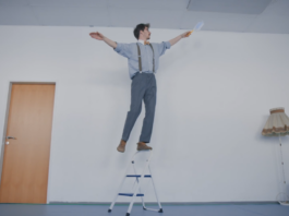 Ein Mann balanciert auf dem Griff einer Trittleiter und hält dabei einen Staubwedel in der Hand.
