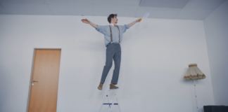 Ein Mann balanciert auf dem Griff einer Trittleiter und hält dabei einen Staubwedel in der Hand.