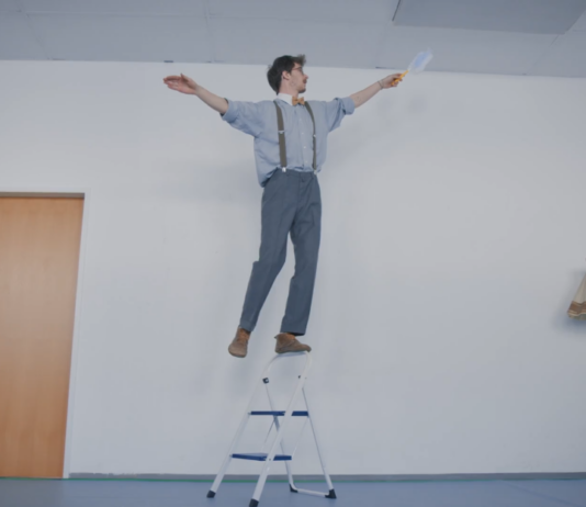 Ein Mann balanciert auf dem Griff einer Trittleiter und hält dabei einen Staubwedel in der Hand.