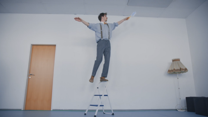 kaudawelsch_tobias mechtler_balanceakt Ein Mann balanciert auf dem Griff einer Trittleiter und hält dabei einen Staubwedel in der Hand.