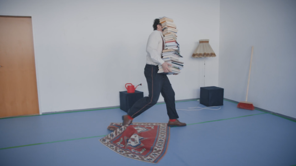 Hubert Wieser trägt einen riesigen Stapel Bücher und stolpert über eine umgeknickte Teppichkante.