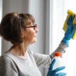 Eine Dame mit einer schicken Bob-Frisur im mittleren Alter putzt ein Fenster. Sie trägt dabei blaue Handschuhe und wischt mit einem gelben Putztuch. In der rechten Hand hält sie eine Flasche eines Reinigungsmittels.