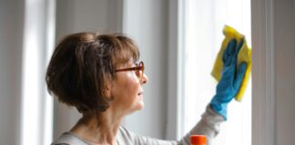 Eine Dame mit einer schicken Bob-Frisur im mittleren Alter putzt ein Fenster. Sie trägt dabei blaue Handschuhe und wischt mit einem gelben Putztuch. In der rechten Hand hält sie eine Flasche eines Reinigungsmittels.
