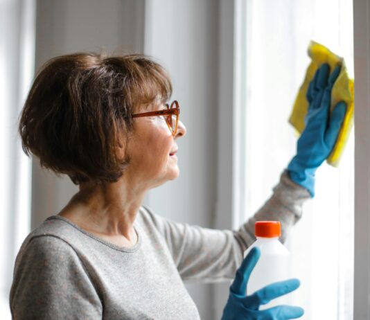 Eine Dame mit einer schicken Bob-Frisur im mittleren Alter putzt ein Fenster. Sie trägt dabei blaue Handschuhe und wischt mit einem gelben Putztuch. In der rechten Hand hält sie eine Flasche eines Reinigungsmittels.