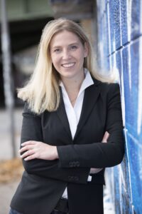 Eine blonde Frau in dunklem Hosenanzug lehnt an einer mit blauem Graffiti besprühter Wand und lächelt mit verschränkten Armen in die Kamera.