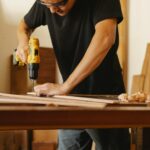 Ein Mann in schwarzem T-Shirt und Schutzbrille steht an einer Werkbank und bohrt in ein Holzbrett.