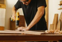 Jährlich verletzen sich beim Heimwerken mehr als 10.000 Menschen Ein Mann in schwarzem T-Shirt und Schutzbrille steht an einer Werkbank und bohrt in ein Holzbrett.