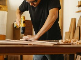 Ein Mann in schwarzem T-Shirt und Schutzbrille steht an einer Werkbank und bohrt in ein Holzbrett.