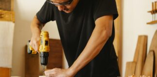 Ein Mann in schwarzem T-Shirt und Schutzbrille steht an einer Werkbank und bohrt in ein Holzbrett.