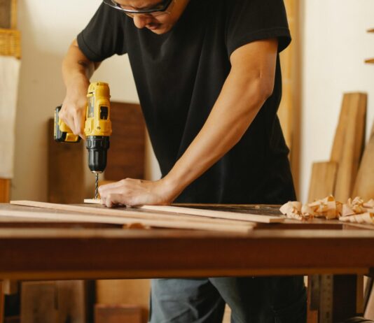 Ein Mann in schwarzem T-Shirt und Schutzbrille steht an einer Werkbank und bohrt in ein Holzbrett.