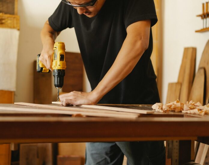 Ein Mann in schwarzem T-Shirt und Schutzbrille steht an einer Werkbank und bohrt in ein Holzbrett.