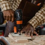 Eine schwarze Person in kariertem Hemd und mit einer ledernen Werkzeug-Schürze bohrt auf einer Werkbank in 2 fixierte Bretter mit einer Bohrmaschine.