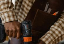 Eine schwarze Person in kariertem Hemd und mit einer ledernen Werkzeug-Schürze bohrt auf einer Werkbank in 2 fixierte Bretter mit einer Bohrmaschine.