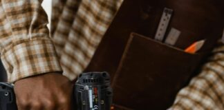 Eine schwarze Person in kariertem Hemd und mit einer ledernen Werkzeug-Schürze bohrt auf einer Werkbank in 2 fixierte Bretter mit einer Bohrmaschine.