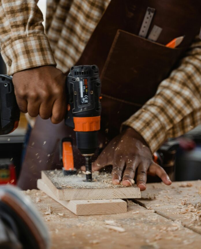 pexels-tima-miroshnichenko-6790048 Eine schwarze Person in kariertem Hemd und mit einer ledernen Werkzeug-Schürze bohrt auf einer Werkbank in 2 fixierte Bretter mit einer Bohrmaschine.