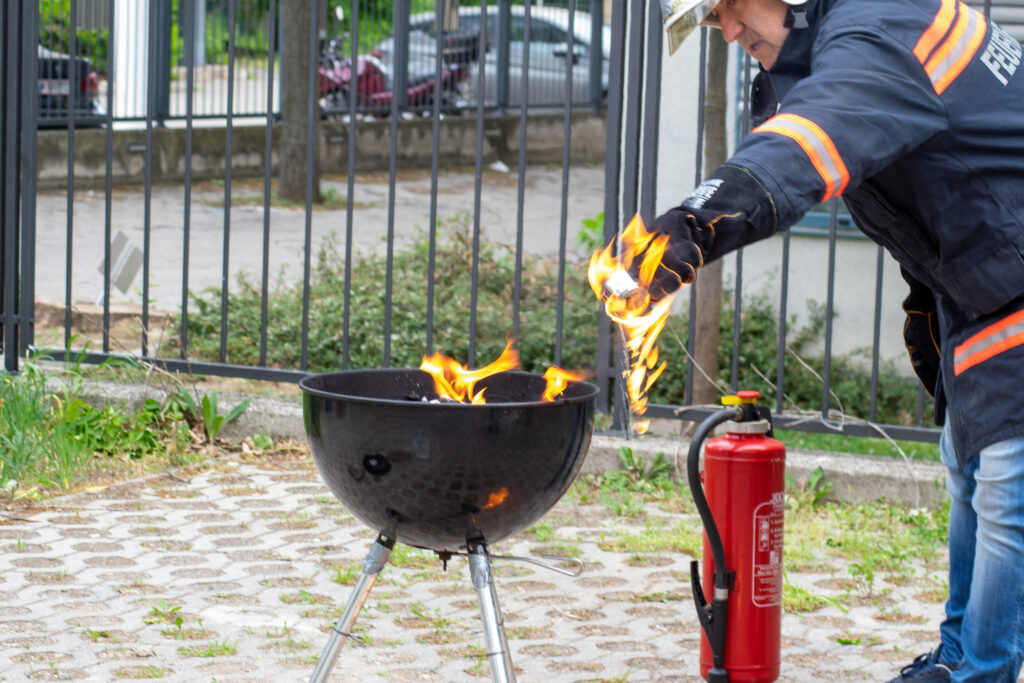 Auf diesem Bild ist ein brennender Grill zu sehen und ein Feuerwehrmann, der ein brennendes Gefäß in der - mit einem feuerfesten Handschuh geschützten - Hand hält. Die Flammen sind auf den Benzin im Gefäß des Eingießers übergesprungen und tropfen nun nach unten auf den Boden. 