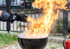 Hier ist ein Griller mit einer Stichflamme zu sehen. Eine in einem feuerfesten Handschuh steckende Hand gießt nur wenig Wasser in die Flammen des Fettbrands und löst damit eine gefährliche Stichflamme aus.