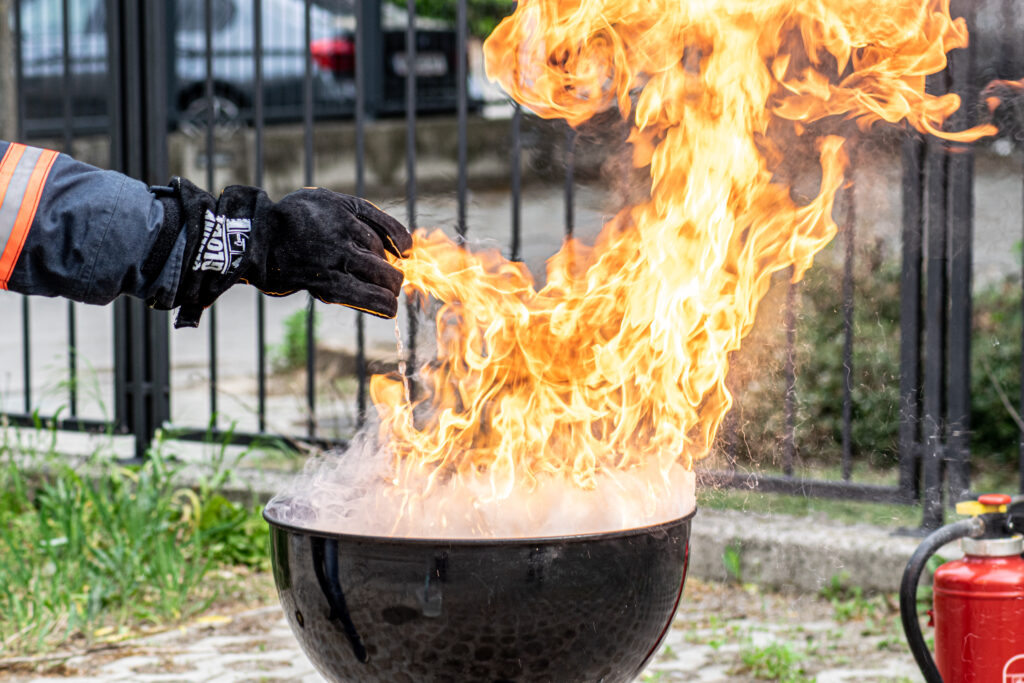 Hier ist ein Griller mit einer Stichflamme zu sehen. Eine in einem feuerfesten Handschuh steckende Hand gießt nur wenig Wasser in die Flammen des Fettbrands und löst damit eine gefährliche Stichflamme aus. 