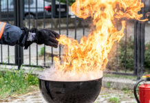Hier ist ein Griller mit einer Stichflamme zu sehen. Eine in einem feuerfesten Handschuh steckende Hand gießt nur wenig Wasser in die Flammen des Fettbrands und löst damit eine gefährliche Stichflamme aus.