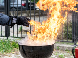 Hier ist ein Griller mit einer Stichflamme zu sehen. Eine in einem feuerfesten Handschuh steckende Hand gießt nur wenig Wasser in die Flammen des Fettbrands und löst damit eine gefährliche Stichflamme aus.