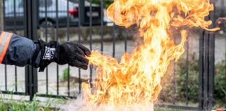Hier ist ein Griller mit einer Stichflamme zu sehen. Eine in einem feuerfesten Handschuh steckende Hand gießt nur wenig Wasser in die Flammen des Fettbrands und löst damit eine gefährliche Stichflamme aus.