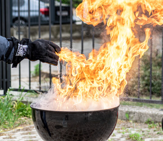 Hier ist ein Griller mit einer Stichflamme zu sehen. Eine in einem feuerfesten Handschuh steckende Hand gießt nur wenig Wasser in die Flammen des Fettbrands und löst damit eine gefährliche Stichflamme aus.
