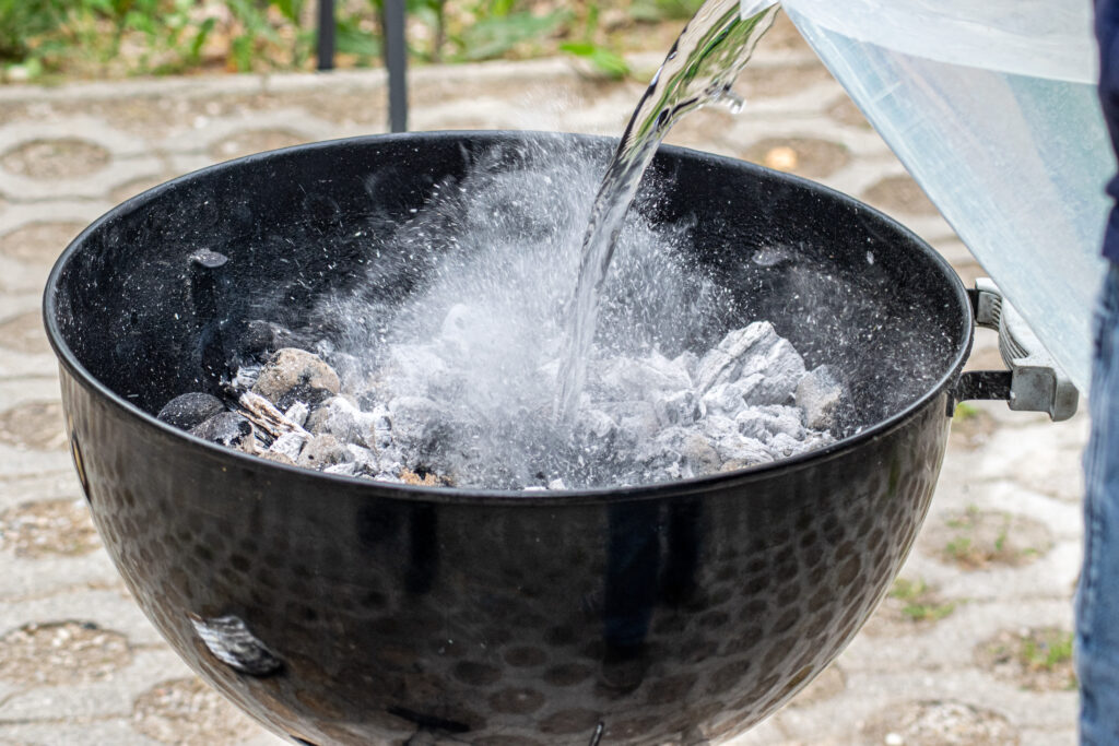 Ein Kugelgrill mit qualmender Kohle. Ein Kübel Wasser wird hinzugeschüttet.