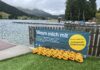Gelbe Schwimmbojen aneinandergereiht am Ufer eines Sees. An dem Zaun vor dem Steg ist ein Banner angebracht mit dem Text: "Nimm mich mit. Boje nehmen - Gegenstände ins Staufach packen. 3x wickeln, verschließen - Hüftgurt umlegen. Schwimmen - zum Ausruhen über die Boje lehnen. Boje zurückbringen. Nie allein schwimmen. Gratisverleih Schwimmboje"