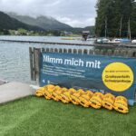 Gelbe Schwimmbojen aneinandergereiht am Ufer eines Sees. An dem Zaun vor dem Steg ist ein Banner angebracht mit dem Text: "Nimm mich mit. Boje nehmen - Gegenstände ins Staufach packen. 3x wickeln, verschließen - Hüftgurt umlegen. Schwimmen - zum Ausruhen über die Boje lehnen. Boje zurückbringen. Nie allein schwimmen. Gratisverleih Schwimmboje"