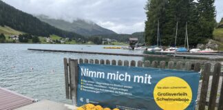 Gelbe Schwimmbojen aneinandergereiht am Ufer eines Sees. An dem Zaun vor dem Steg ist ein Banner angebracht mit dem Text: "Nimm mich mit. Boje nehmen - Gegenstände ins Staufach packen. 3x wickeln, verschließen - Hüftgurt umlegen. Schwimmen - zum Ausruhen über die Boje lehnen. Boje zurückbringen. Nie allein schwimmen. Gratisverleih Schwimmboje"