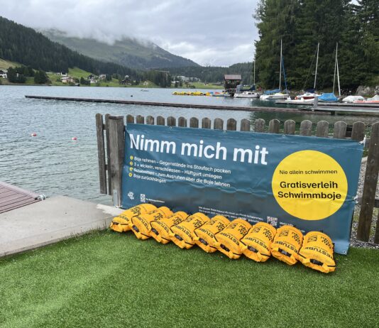 Gelbe Schwimmbojen aneinandergereiht am Ufer eines Sees. An dem Zaun vor dem Steg ist ein Banner angebracht mit dem Text: "Nimm mich mit. Boje nehmen - Gegenstände ins Staufach packen. 3x wickeln, verschließen - Hüftgurt umlegen. Schwimmen - zum Ausruhen über die Boje lehnen. Boje zurückbringen. Nie allein schwimmen. Gratisverleih Schwimmboje"