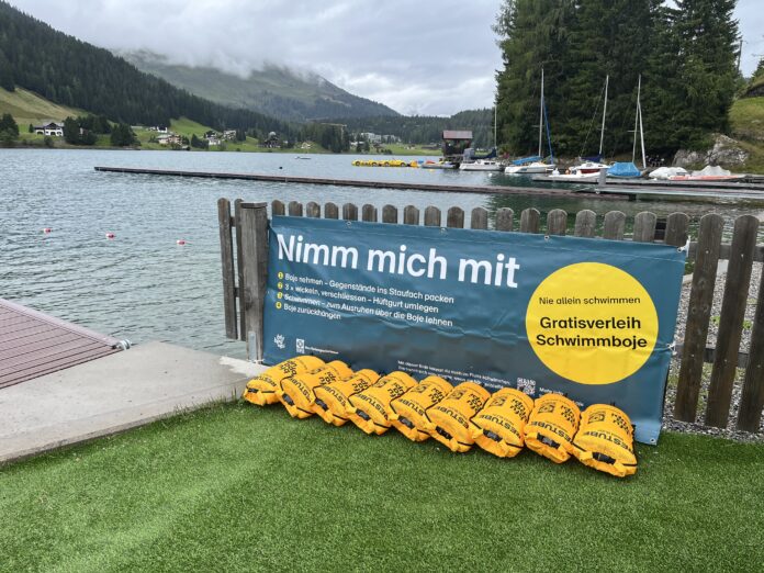 2025-08-12_BojenverleihstationDavosersee_Quelle-GemeindeDavos Gelbe Schwimmbojen aneinandergereiht am Ufer eines Sees. An dem Zaun vor dem Steg ist ein Banner angebracht mit dem Text: "Nimm mich mit. Boje nehmen - Gegenstände ins Staufach packen. 3x wickeln, verschließen - Hüftgurt umlegen. Schwimmen - zum Ausruhen über die Boje lehnen. Boje zurückbringen. Nie allein schwimmen. Gratisverleih Schwimmboje"