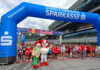 Kinder und Jugendliche stehen beim Businesslauf 2026 am Red Bull Ring unter einem blauen Startbogen der Steiermärkischen Sparkasse, davor zwei Maskottchen.