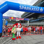 Kinder und Jugendliche stehen beim Businesslauf 2026 am Red Bull Ring unter einem blauen Startbogen der Steiermärkischen Sparkasse, davor zwei Maskottchen.