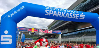 Kinder und Jugendliche stehen beim Businesslauf 2026 am Red Bull Ring unter einem blauen Startbogen der Steiermärkischen Sparkasse, davor zwei Maskottchen.
