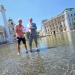 Umweltmediziner und Ökologe Prof. Dr. Hans-Peter Hutter von der MedUni Wien und Dr. Armin Kaltenegger, Leiter des Fachbereichs Eigentumsschutz im KFV stehen am Karlsplatz im künstlich angelegten Teich mit kurzer Hose bzw. aufgekrempelter Anzugshose. Sie halten ein Schild mit "40°C" hoch. Neben ihnen im Wasser schwimmt eine umgekippte Badeente. Im Hintergrund sind die Karlskirche, Gebäude und Bäume zu sehen.
