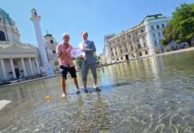 Umweltmediziner und Ökologe Prof. Dr. Hans-Peter Hutter von der MedUni Wien und Dr. Armin Kaltenegger, Leiter des Fachbereichs Eigentumsschutz im KFV stehen am Karlsplatz im künstlich angelegten Teich mit kurzer Hose bzw. aufgekrempelter Anzugshose. Sie halten ein Schild mit "40°C" hoch. Neben ihnen im Wasser schwimmt eine umgekippte Badeente. Im Hintergrund sind die Karlskirche, Gebäude und Bäume zu sehen.