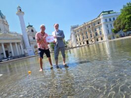 Umweltmediziner und Ökologe Prof. Dr. Hans-Peter Hutter von der MedUni Wien und Dr. Armin Kaltenegger, Leiter des Fachbereichs Eigentumsschutz im KFV stehen am Karlsplatz im künstlich angelegten Teich mit kurzer Hose bzw. aufgekrempelter Anzugshose. Sie halten ein Schild mit "40°C" hoch. Neben ihnen im Wasser schwimmt eine umgekippte Badeente. Im Hintergrund sind die Karlskirche, Gebäude und Bäume zu sehen.