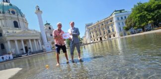 Umweltmediziner und Ökologe Prof. Dr. Hans-Peter Hutter von der MedUni Wien und Dr. Armin Kaltenegger, Leiter des Fachbereichs Eigentumsschutz im KFV stehen am Karlsplatz im künstlich angelegten Teich mit kurzer Hose bzw. aufgekrempelter Anzugshose. Sie halten ein Schild mit "40°C" hoch. Neben ihnen im Wasser schwimmt eine umgekippte Badeente. Im Hintergrund sind die Karlskirche, Gebäude und Bäume zu sehen.