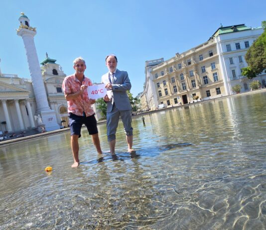 Umweltmediziner und Ökologe Prof. Dr. Hans-Peter Hutter von der MedUni Wien und Dr. Armin Kaltenegger, Leiter des Fachbereichs Eigentumsschutz im KFV stehen am Karlsplatz im künstlich angelegten Teich mit kurzer Hose bzw. aufgekrempelter Anzugshose. Sie halten ein Schild mit "40°C" hoch. Neben ihnen im Wasser schwimmt eine umgekippte Badeente. Im Hintergrund sind die Karlskirche, Gebäude und Bäume zu sehen.