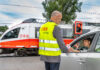 Ein Mann in ÖBB-Sicherheitsweste vor einem Bahnübergang reicht einer Frau im Auto einen Informationsflyer. Das Auto steht an einer roten Ampel an einem Bahnübergang. Ein ÖBB-Zug fährt gerade durch.