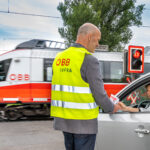 Ein Mann in ÖBB-Sicherheitsweste vor einem Bahnübergang reicht einer Frau im Auto einen Informationsflyer. Das Auto steht an einer roten Ampel an einem Bahnübergang. Ein ÖBB-Zug fährt gerade durch.