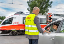Ein Mann in ÖBB-Sicherheitsweste vor einem Bahnübergang reicht einer Frau im Auto einen Informationsflyer. Das Auto steht an einer roten Ampel an einem Bahnübergang. Ein ÖBB-Zug fährt gerade durch.